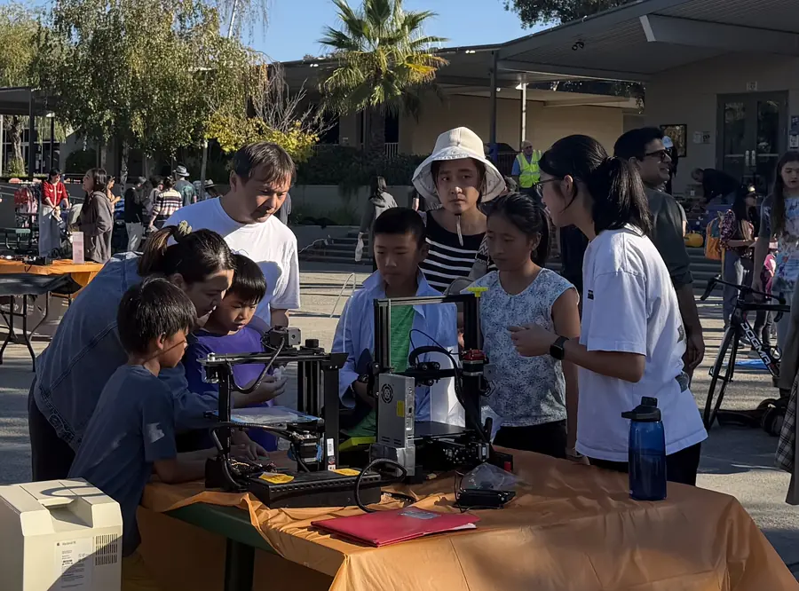 A dense knot of kids and parents crowds around a running 3D printer at a maker booth, a student volunteer in a team shirt explaining the print while a vintage Macintosh sits at the table's edge.