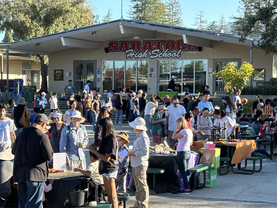 A packed courtyard in warm late-afternoon light: families in sun hats crowd activity tables and green picnic benches beneath the big SARATOGA HIGH SCHOOL letters.