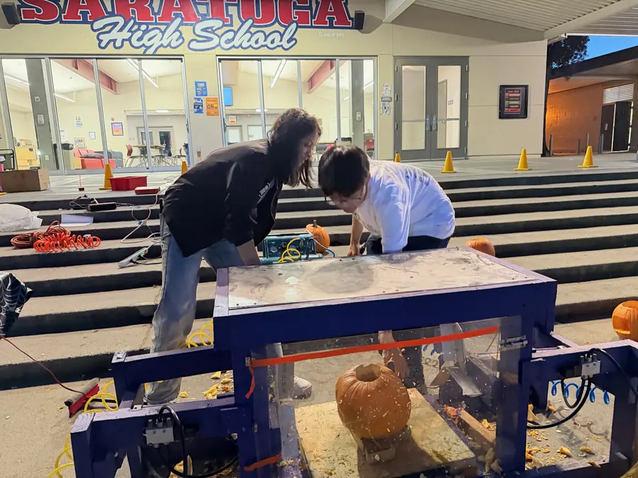 Two students lean over the purple pumpkin-smashing machine at dusk, looking through its pulp-spattered acrylic guard at a pumpkin on the crush plate.