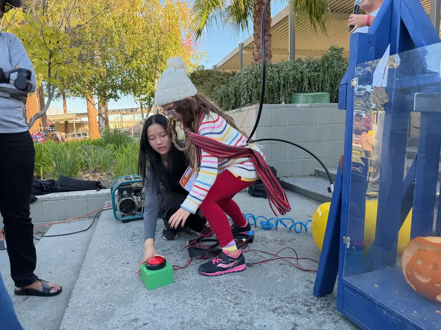 A little girl in a pompom beanie and rainbow-striped sweater lunges to slam the big red arcade button on its green box while a student volunteer crouches beside her and the carved jack-o-lantern waits inside the blue smasher.