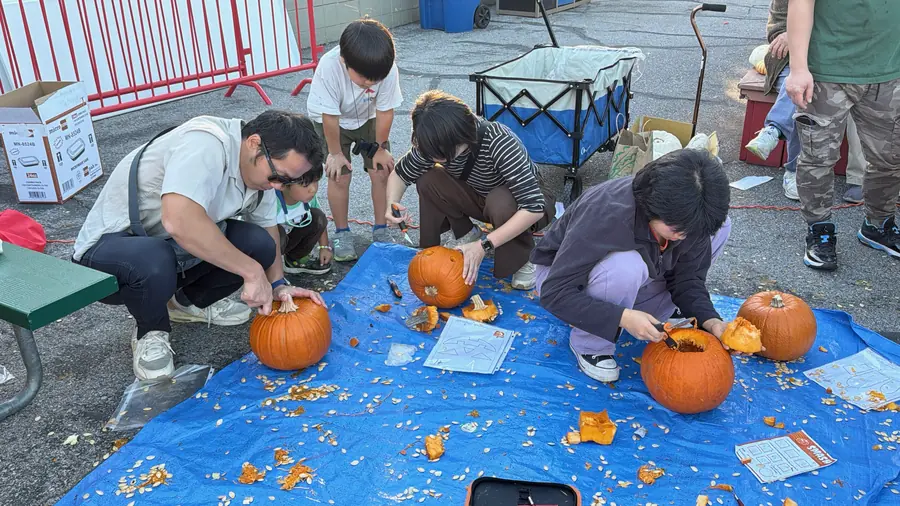Wide, clean view of families carving pumpkins on a blue tarp, three groups working scalpels into jack-o-lantern faces with stencil sheets beside them.