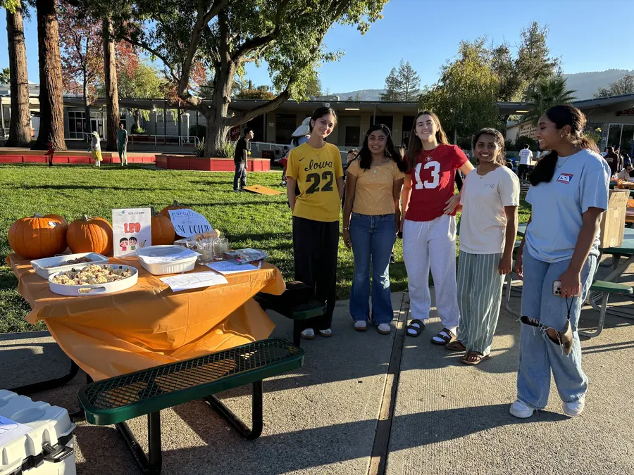 Five smiling high school club members pose beside their orange-draped bake sale table set with pumpkins, LEO club signage and trays of treats on the lawn.