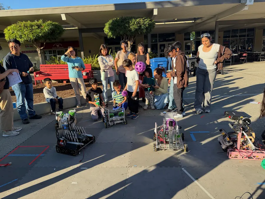 Kids kneel on the pavement driving competition robots with game controllers while a purple game ball sits mid-play and a semicircle of parents watches.