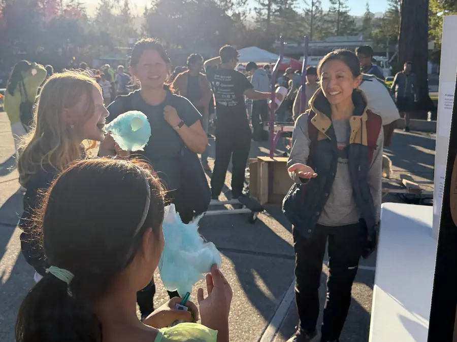 A girl bites into a cloud of blue cotton candy while two smiling women look on in backlit late-afternoon sun, another kid in the foreground holding up her own cone.