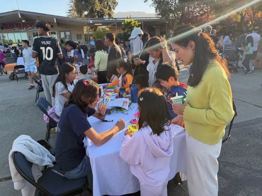 A packed craft and science table with young children folding paper creations while a student volunteer leans in to help.