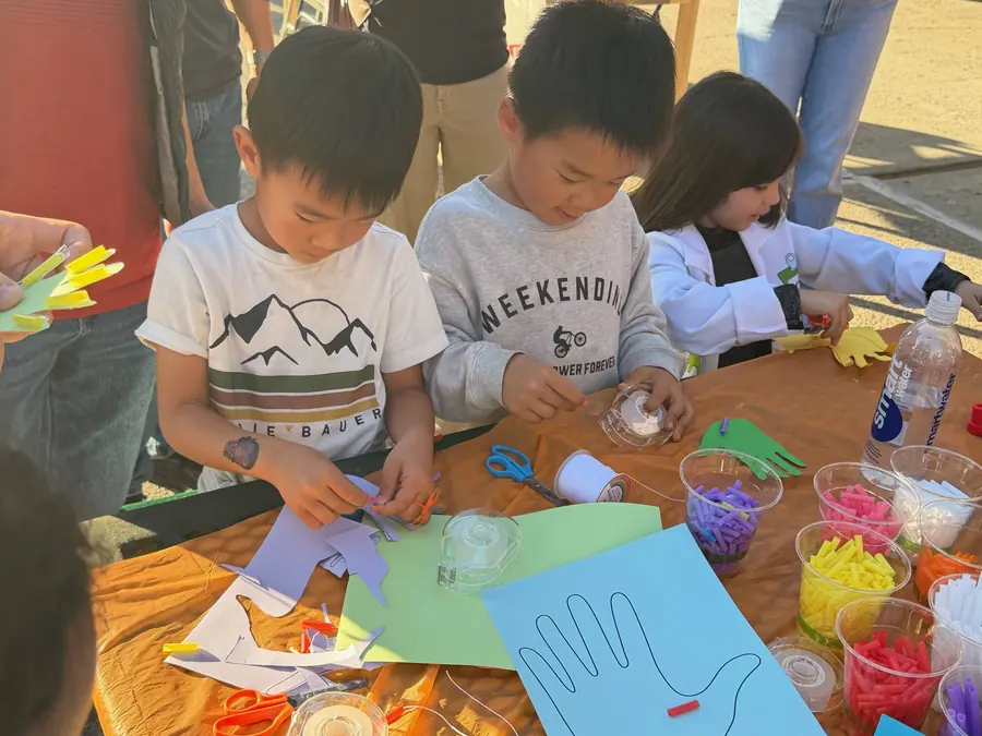 Three young kids in a row lean over the craft table in warm late light, tongue-out concentration as they cut and tape foam strips onto their paper articulated hands.