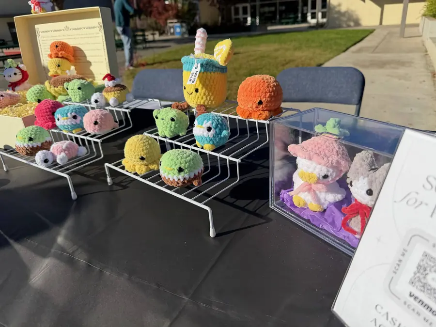 A club booth table lined with dozens of handmade crochet plushies — octopuses, turtles, frogs, a boba tea and a bunny in a display case — glowing in low golden sunlight.