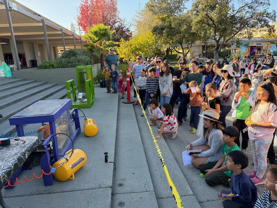 A big crowd of families packs the amphitheater steps behind yellow caution tape, watching the purple pneumatic pumpkin-smashing machine with its yellow air tanks primed on the concrete stage.