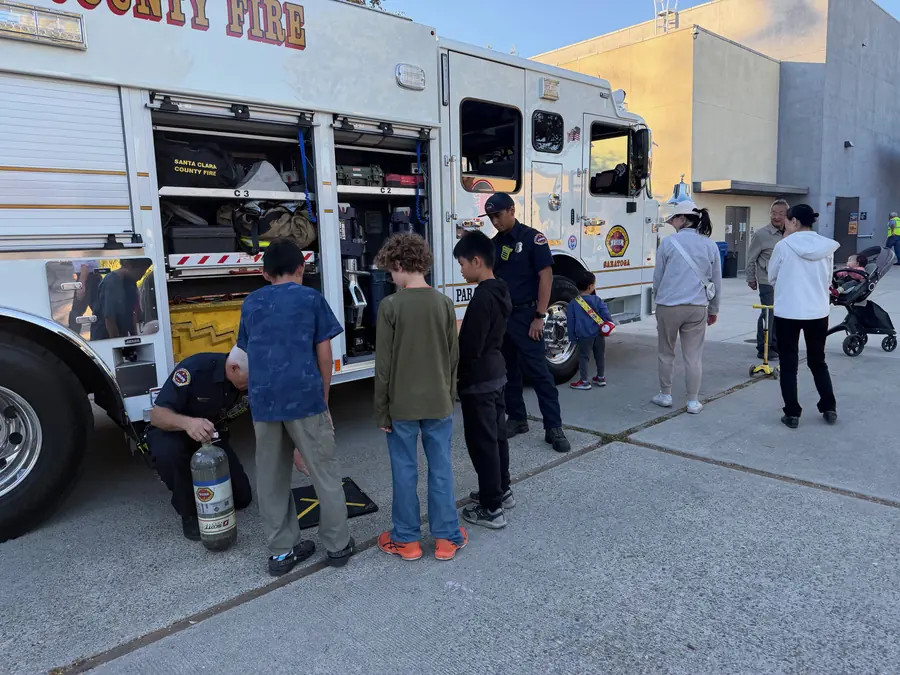 A firefighter kneels to hand a breathing-apparatus air bottle to a line of kids at the open side of the Saratoga fire engine, another firefighter standing with a small child beside the cab.