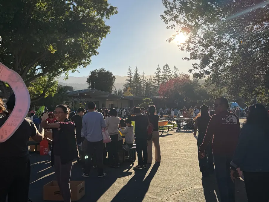 Wide golden-hour view across the packed school courtyard, sun flaring through the oak canopy over a crowd of families at booths with the hills behind the Saratoga High School building.