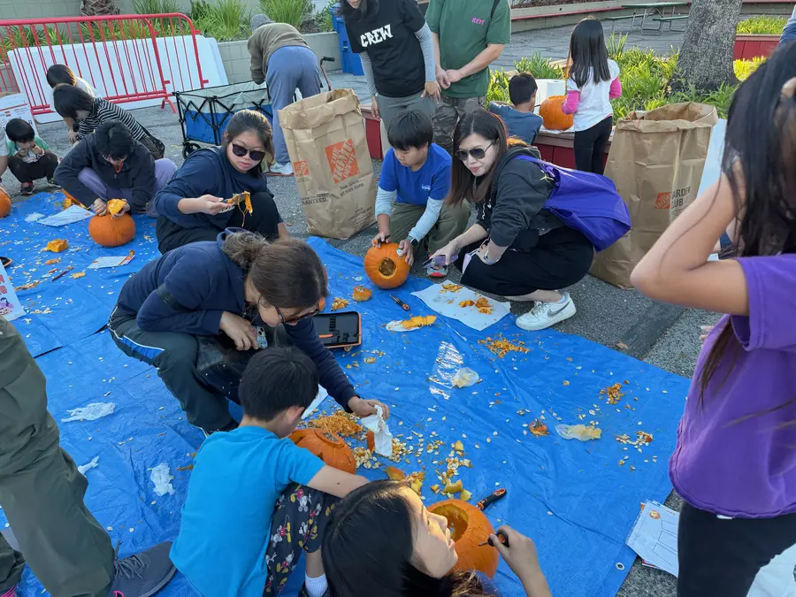 A crowded blue tarp of pumpkin carving — parents and kids kneeling over gutted pumpkins, seeds and pulp everywhere, Home Depot bags staged for compost.