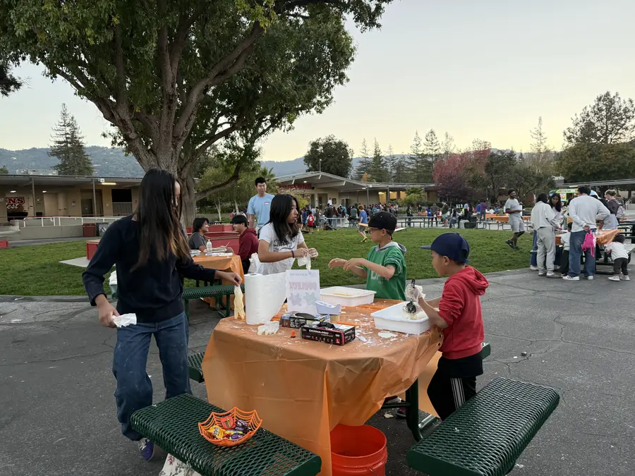 Two boys in ball caps plunge gloved hands into white bins of oobleck at an orange-draped picnic table while student volunteers coach them, the hills going gold behind the courtyard.