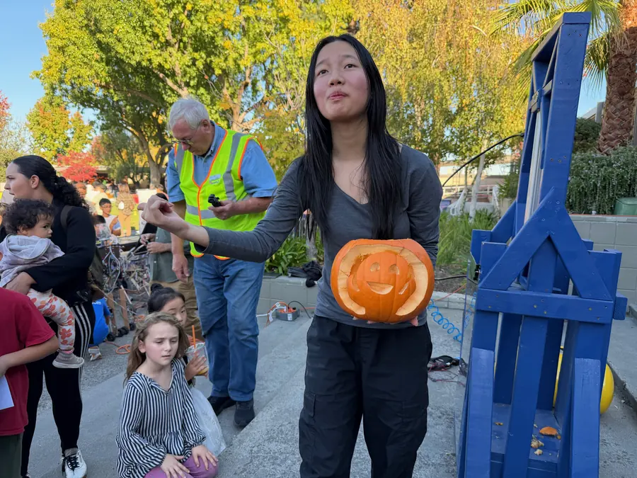 A teen volunteer holds a carved smiley-face jack-o-lantern out toward the blue guillotine smasher, backlit autumn foliage glowing gold behind her.