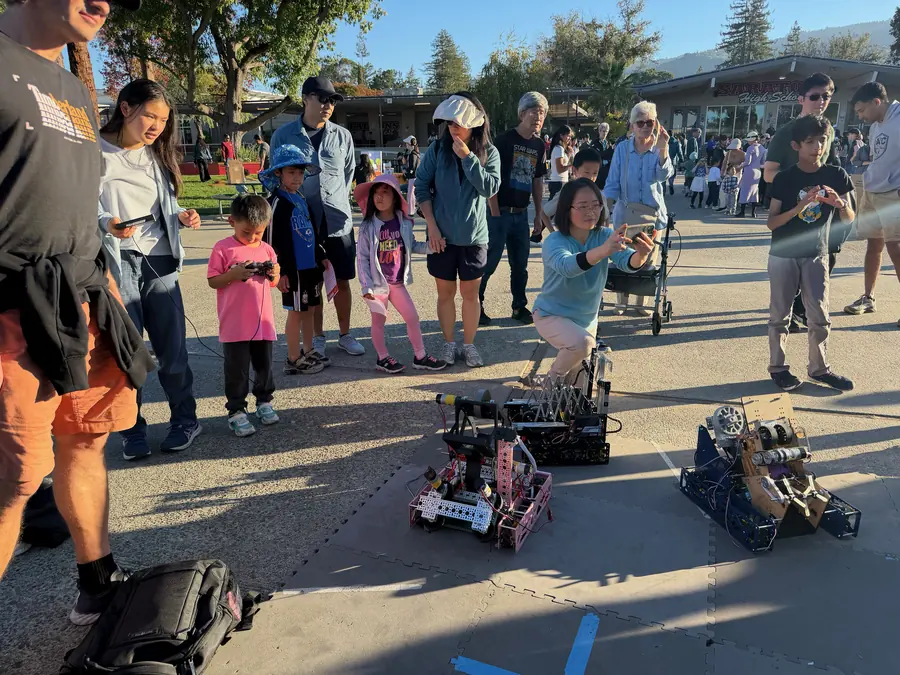 A small boy in a pink shirt drives a robot with a controller while a woman crouches to film three FTC robots on the mat, a whole arc of families watching in clean late-day light with the Saratoga High sign behind.
