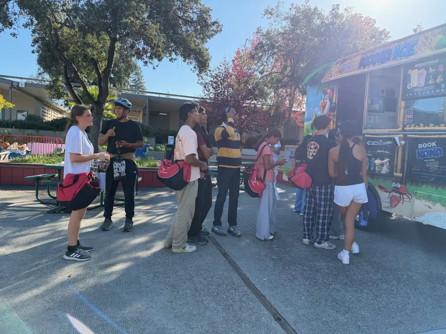A line of teenagers with red gear bags queues at the bright yellow Kona Ice shaved-ice truck in warm late-afternoon light under the courtyard trees.