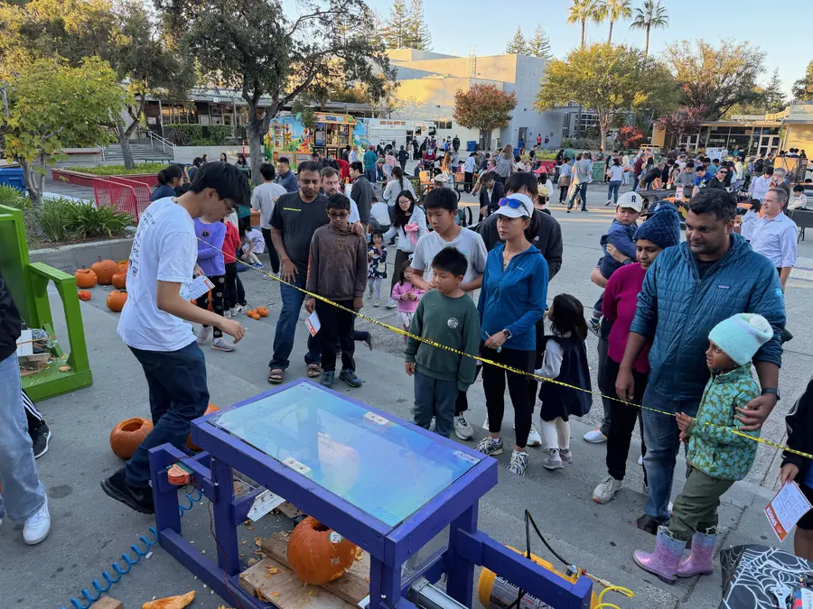 A student operator leans over the purple pumpkin smasher with a pumpkin loaded under the press and a long line waiting behind the tape.