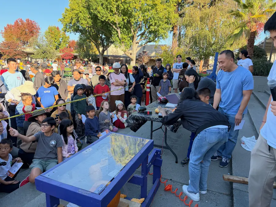 A packed ring of kids sits on the pavement behind caution tape watching a student and an adult volunteer load a pumpkin into the blue acrylic-windowed smashing machine, autumn trees glowing behind the crowd.