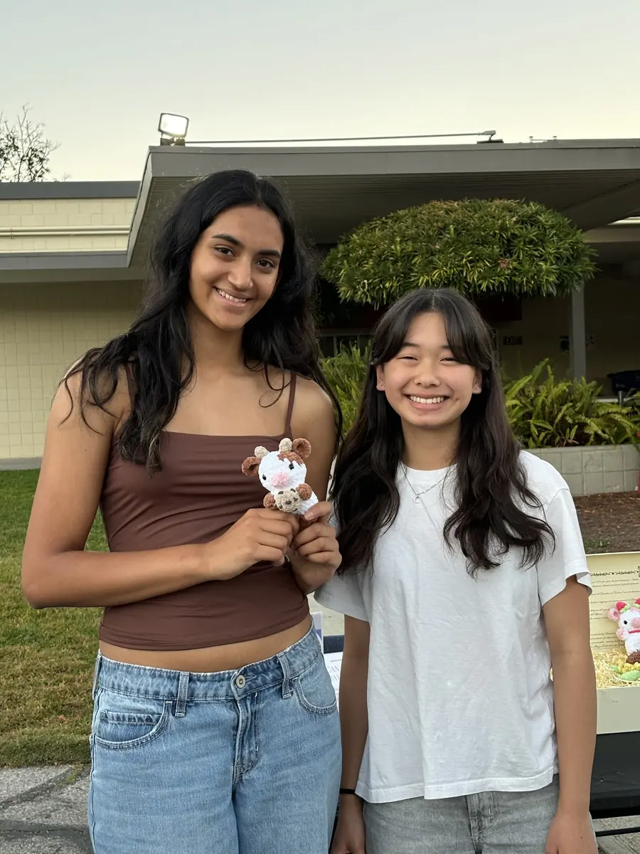 Two student makers grin shoulder to shoulder in warm evening light, holding up a tiny crochet cow they made at their booth.