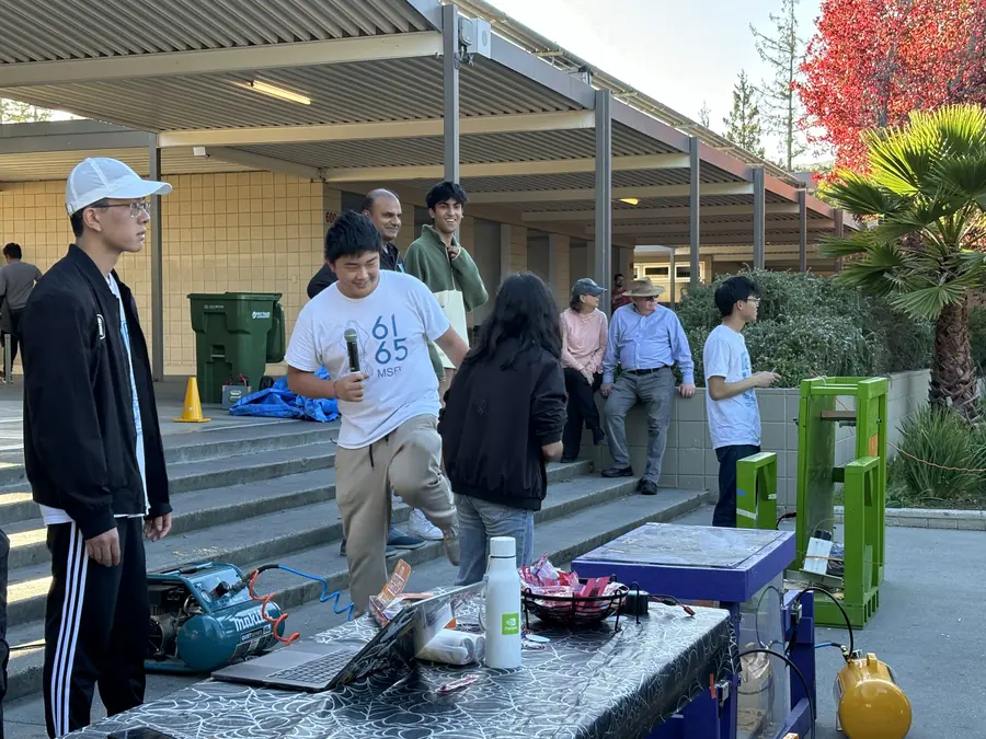 A grinning MSET 6165 student in team shirt strides with a microphone beside the blue pumpkin-smasher rig and its green hydraulic press, air compressor and laptop on the spider-web-cloth table in front of him.