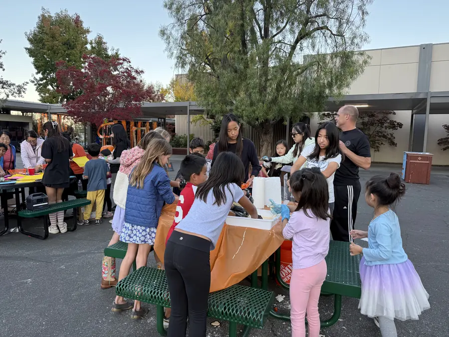 A ring of kids leans over a picnic table plunging gloved hands into a tub of oobleck while teen volunteers manage the mess, autumn maples and the campus roofline behind them.