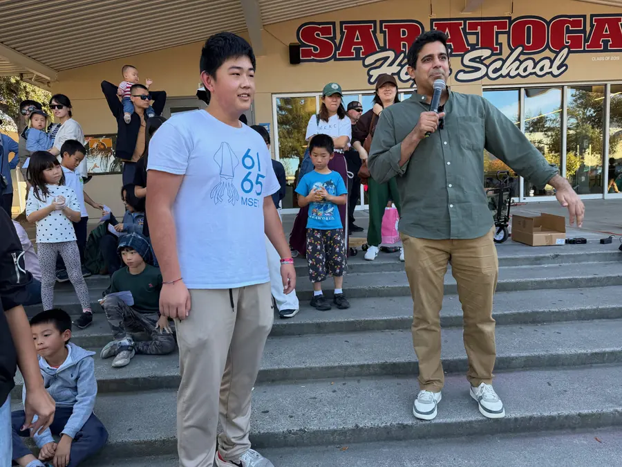 A student from MSET 6165 stands with the event emcee on the school steps under the SARATOGA HIGH SCHOOL sign, a crowd of seated children in the foreground.