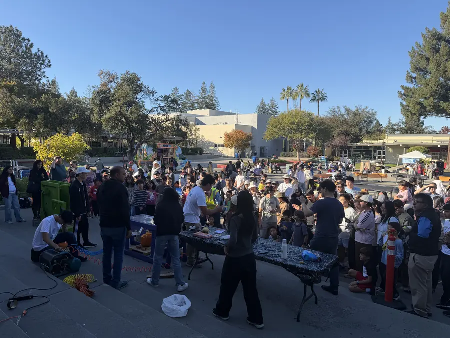 A high wide shot over the whole SMASH fair — a big crowd packed around the pumpkin-smashing machine on the courtyard steps, booths and canopies stretching back across campus under a clear blue sky.