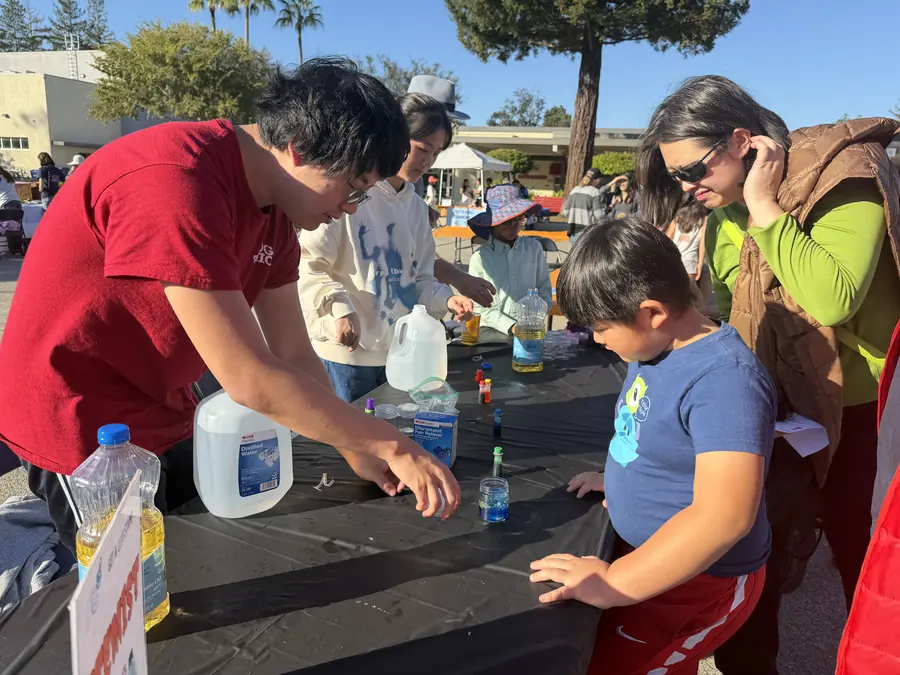 A student in a red Saratoga Physics shirt runs a hands-on demo — oil, water and effervescent tablets in jars — while a young boy leans in over the table and his mother watches under bright blue afternoon sky.