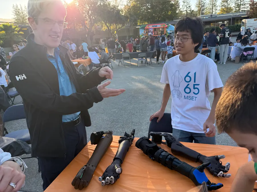 A man demonstrates finger articulation with his own hand while an MSET 6165 student looks on, four robotic prosthetic arms laid out across the orange table between them.