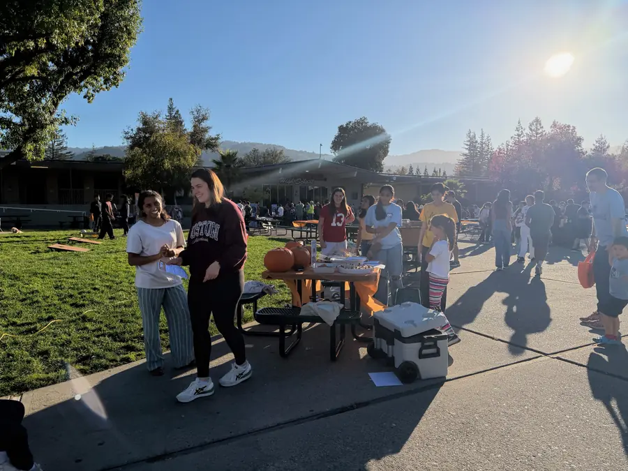 Two teen volunteers walk away from the pumpkin-pie table laughing, backlit by the low sun over the hills with the busy courtyard stretching behind them.