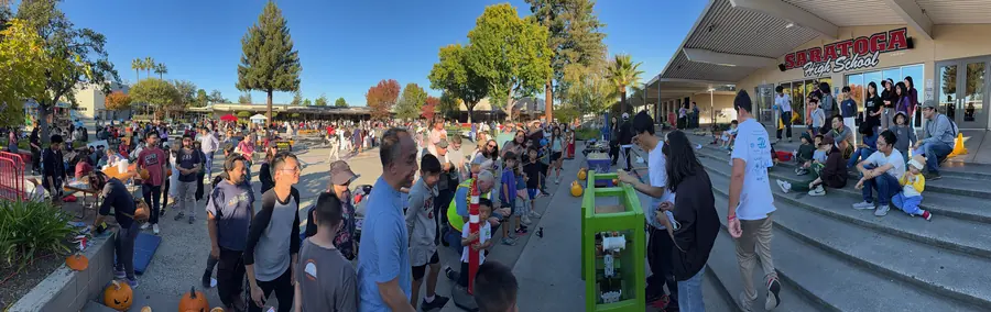 Ultra-wide panorama of the packed Saratoga High courtyard at golden hour, the green pumpkin-smashing machine and the school sign anchoring the right side.