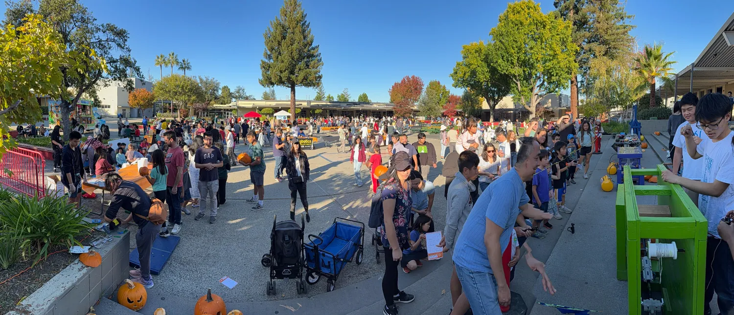 Panorama across the whole Saratoga High courtyard at peak attendance, carving tables and jack-o’-lanterns in the foreground and hundreds of visitors filling the plaza.