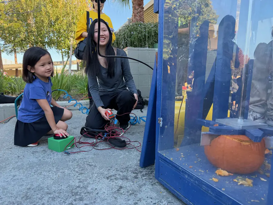 A young girl crouches with her hand on a large red button mounted to a green box, grinning, while a student volunteer steadies the cable beside her and a pumpkin waits inside the machine’s clear chamber.