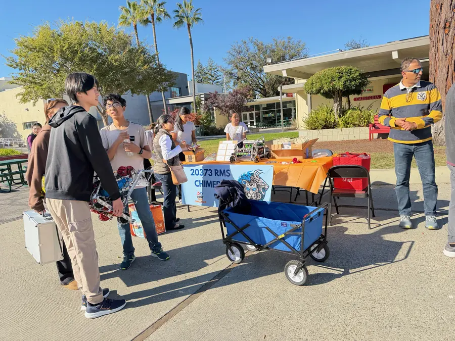 Visitors and students gather around FTC team 20373 RMS Chimera's orange-draped booth, robots lined up on the table under palm trees and blue sky.