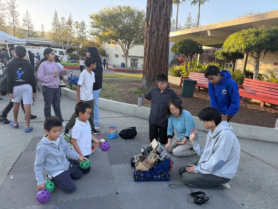 Two little boys clutching purple and green game balls kneel on the foam mat watching an FTC robot while high-schoolers and a parent crouch around it, explaining, in warm late-afternoon light under a redwood.