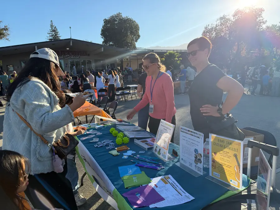 Two smiling library staffers greet a visitor at an information table spread with pens, flyers and green stress balls, sun flaring over the courtyard behind them.