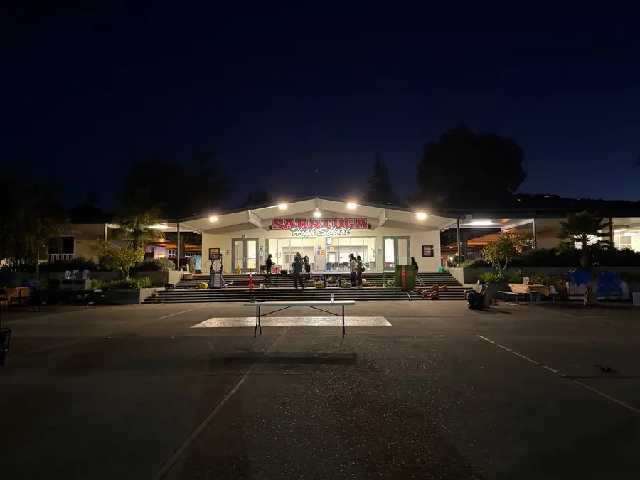 The Saratoga High courtyard after dark, the lit school marquee glowing at the far end of an empty asphalt plaza with a lone folding table in the foreground and a handful of people packing up on the steps.