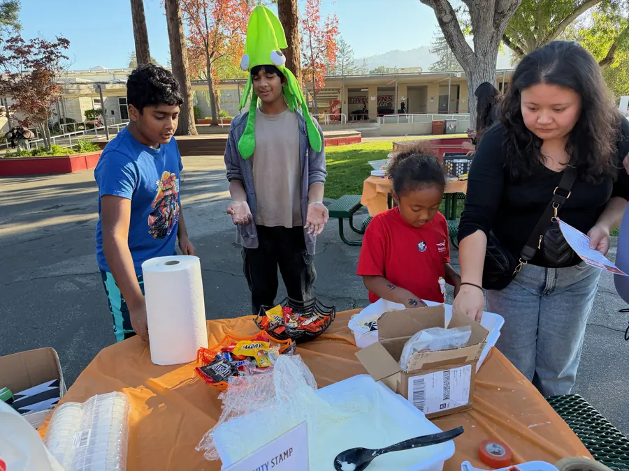 Kids crowd an orange-clothed activity table stocked with candy and slime supplies, one boy in an inflatable green squid hat holding out his goo-covered hands.