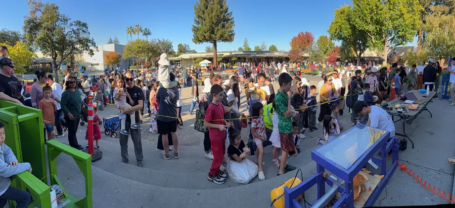 Sweeping panorama across the Saratoga High courtyard showing both smashing machines, the roped-off crowd, and rows of booths and visitors stretching back toward the buildings under a clear sky.