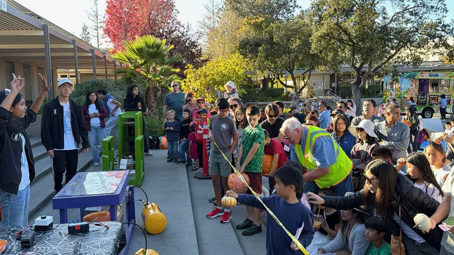 A student throws her arms up in celebration at the smasher controls while kids clutching pumpkins queue along the tape and a volunteer in a hi-vis vest keeps the line moving.