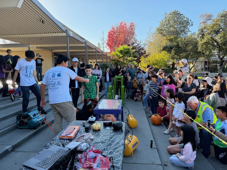 A student operator gestures a boy in a green shirt up to the smasher's control table, pumpkins and prize bags laid out in front, as a wide arc of families and phone cameras watches from the steps.