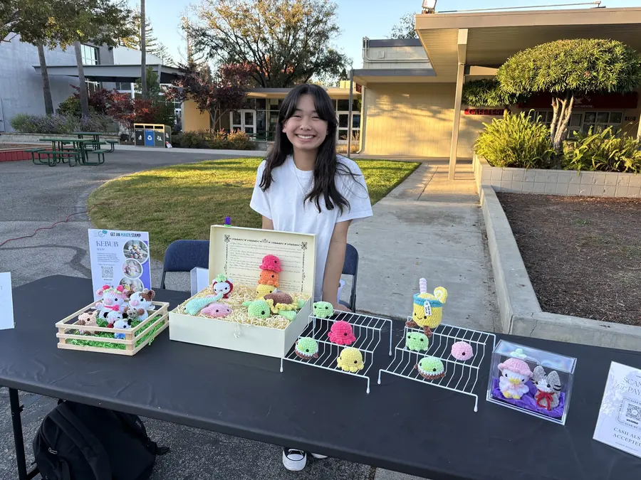 A beaming student vendor stands behind her tidy black booth table displaying rows of tiny handmade crochet animals and boba charms in the low golden light.