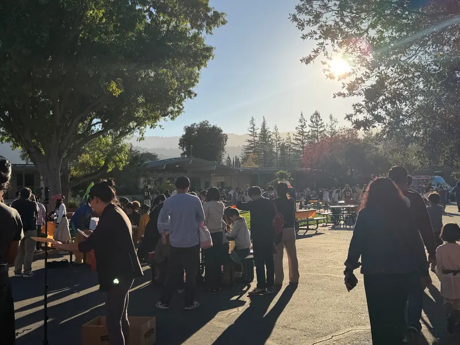 Golden-hour wide shot of the courtyard with sun bursting through the trees, long shadows across the blacktop and a dense line of families browsing booths toward the school building and hills.