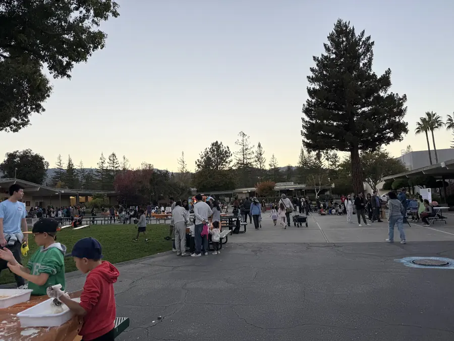 Sweeping sunset view down the whole courtyard: a long spine of activity tables and hundreds of people stretching toward the buildings, a lone redwood spiking into an enormous open sky, oobleck kids at the near edge.