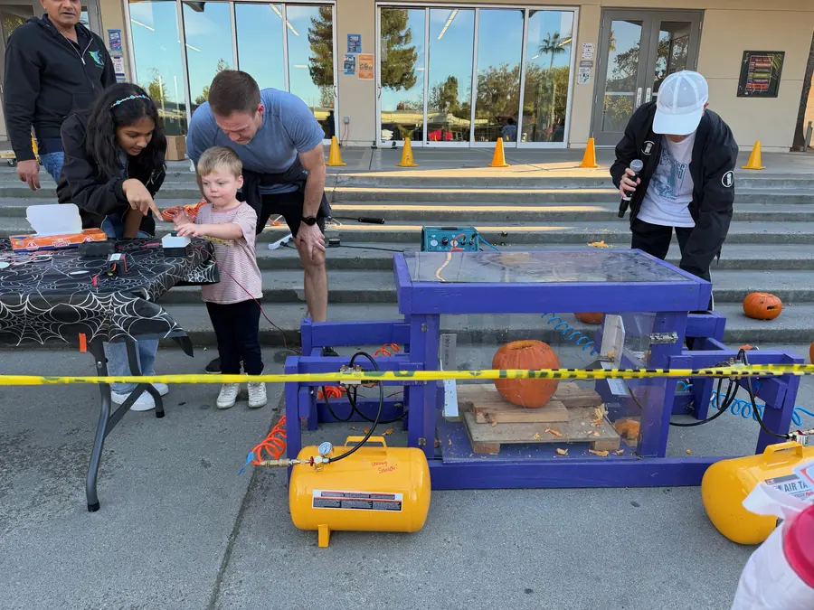 A small blond boy, guided by his dad and a student volunteer, reaches for the firing button of the purple pneumatic pumpkin-smashing machine with a whole pumpkin waiting inside the clear box.