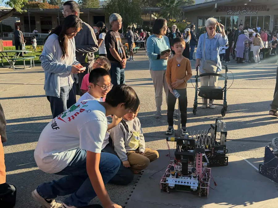 Two MSET students crouch on the blacktop tuning a pink FTC robot while a multigenerational crowd — kids, parents, a grandmother with a walker — watches from behind, the Saratoga High School sign glowing in the background.