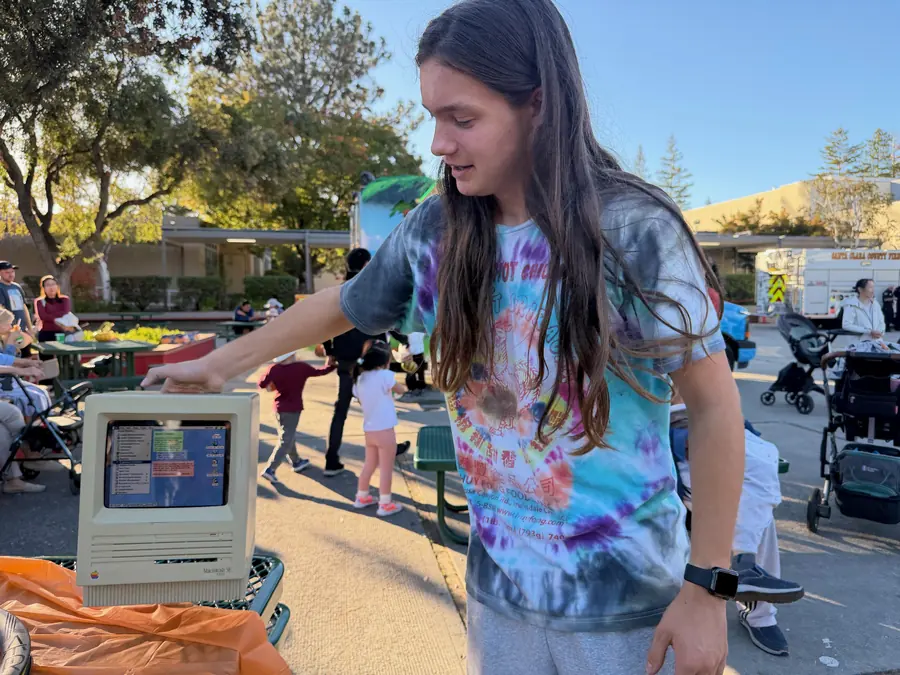 A long-haired teen in a vivid tie-dye shirt rests a hand atop a vintage Macintosh SE running a classic Mac desktop, grinning down at it in warm side light.