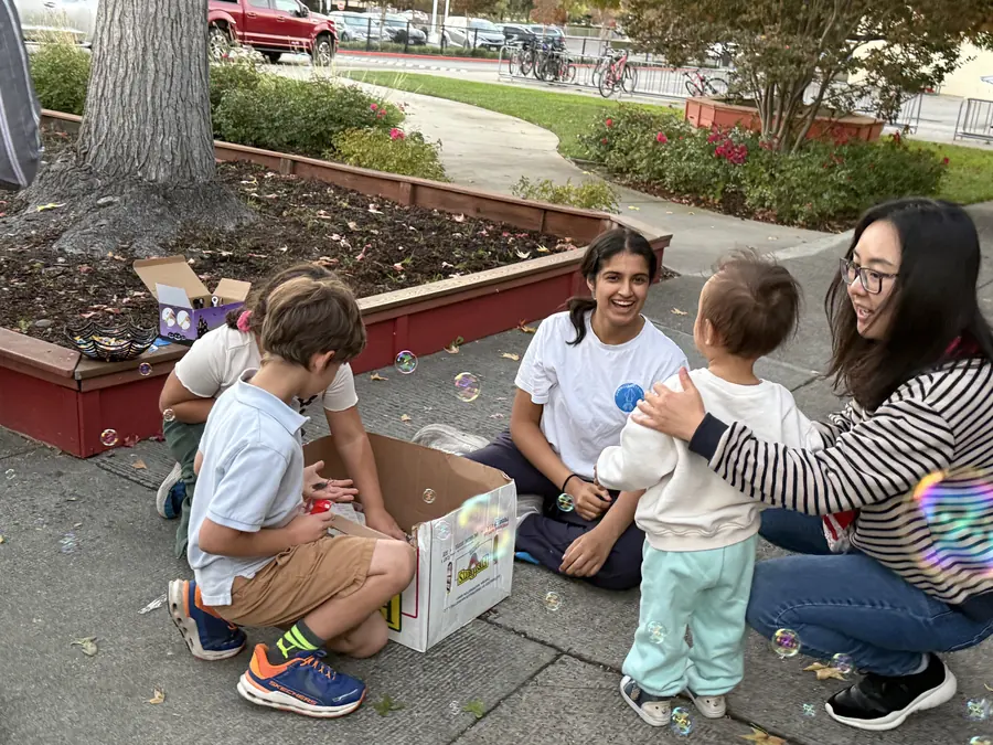 A student volunteer in a white MSET shirt sits on the pavement laughing with a toddler while a mom kneels alongside and soap bubbles drift through the whole frame.
