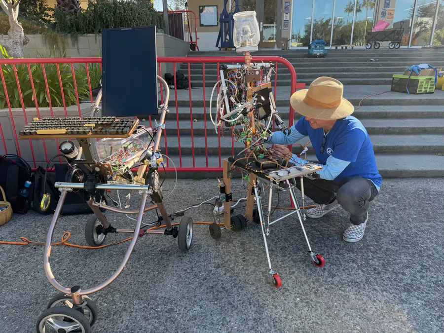 A maker in a straw sun hat crouches on the pavement adjusting two humanoid sculptures built on repurposed walker frames, all copper tubing, exposed circuit boards and looping wire.
