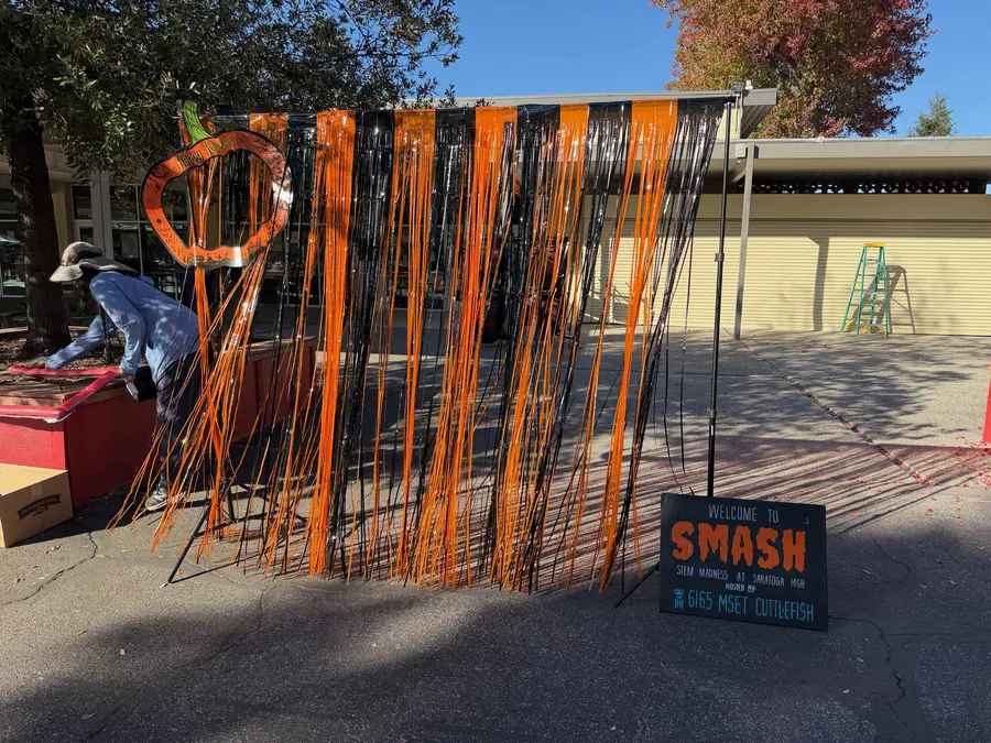 A chalkboard sign reading “Welcome to SMASH — STEM Madness at Saratoga High, hosted by 6165 MSET Cuttlefish” beside an orange-and-black streamer curtain.
